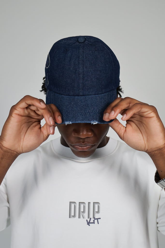 Model wearing dark denim baseball cap with flame embroidery and "DESIGNER DETOUR" patch, paired with white streetwear sweatshirt featuring bold "DRIP KIT" logo – modern urban fashion look, unisex edgy cap styling, premium streetwear brand photography.

 baseball millennials couples streetwear style fashion sustainable eco-friendly vacation daily essentials Gen Z cap hat bold statement luxury cool summer clothing trend outfit OOTD Instagram designer dripkit eco-fashion streetwear 
